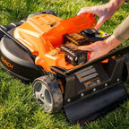 Person inserting a battery into an orange and black lawn mower on grass
