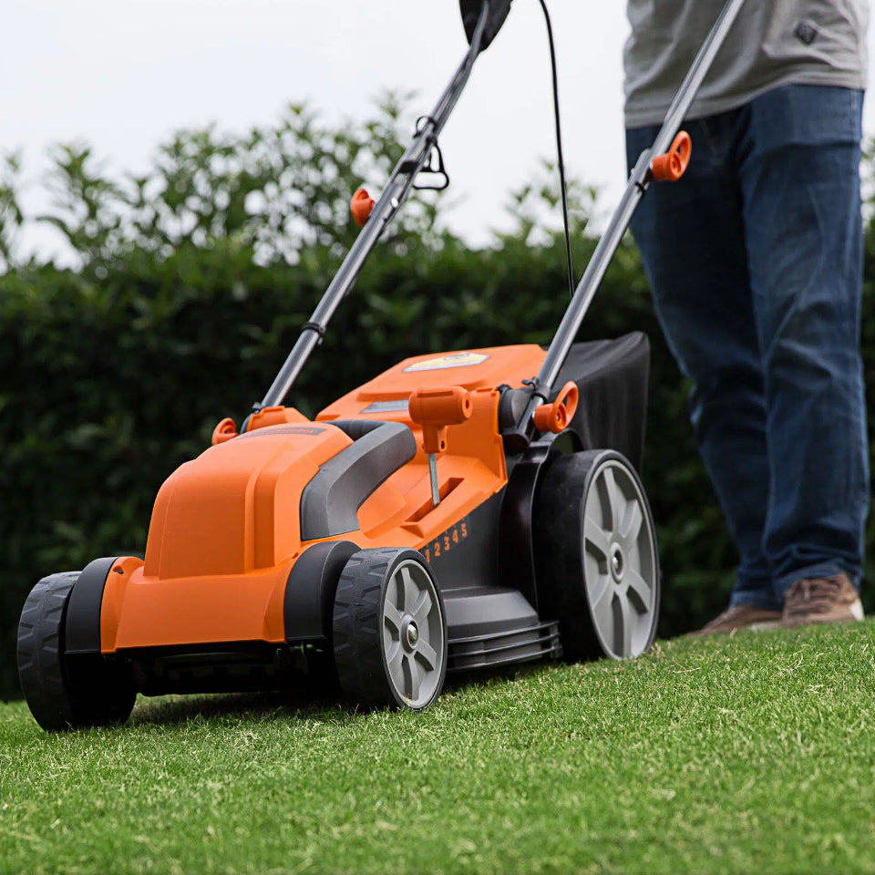Person using an orange and black lawn mower on a grassy area with greenery in the background.