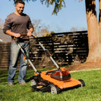 Man using a lawn mower on a grassy area with a wooden fence and tree in the background.