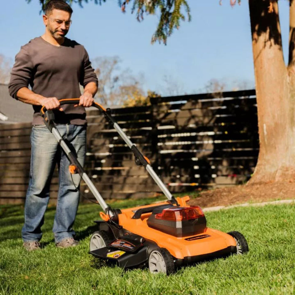 Man using a lawn mower on a grassy area with a wooden fence and tree in the background.