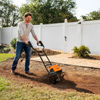 Man using a tiller on a garden bed with a white fence and trees in the background