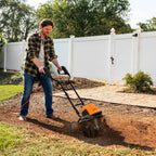 Man using a tiller in a garden with a white fence and trees in the background