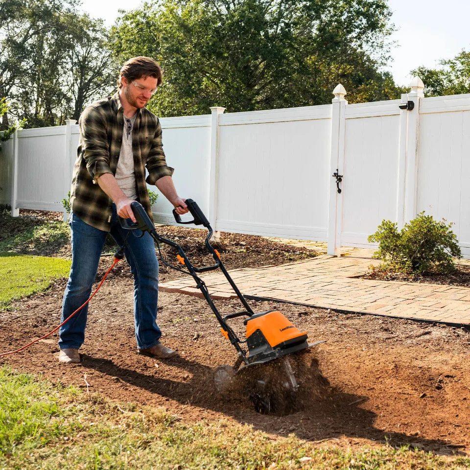 Man using a tiller in a garden with a white fence and trees in the background