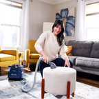 Woman cleaning a white ottoman with a vacuum cleaner in a living room.