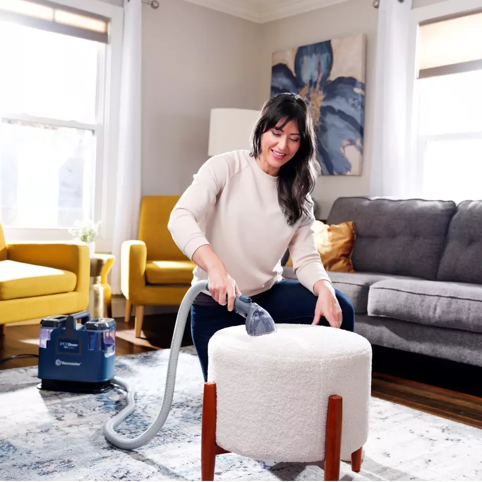 Woman cleaning a white ottoman with a vacuum cleaner in a living room.