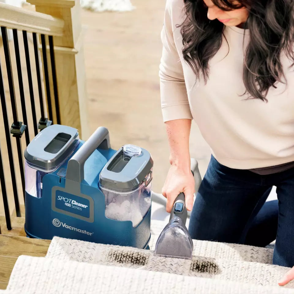 Person using a Vacmaster Spot Cleaner on a rug with a blue cleaning machine in the foreground.