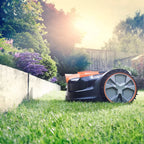 Robotic lawn mower cutting grass in a garden with trees and a fence in the background.